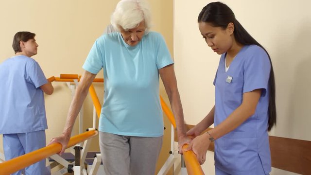 Tracking shot of young male and female physiotherapy specialists in scrubs helping elderly people learning to walk after stroke or injury
