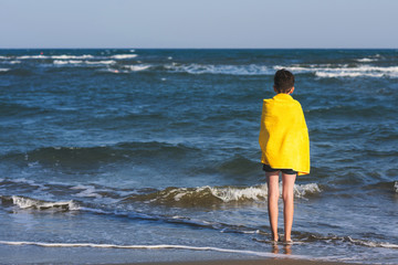 Back view on the boy standing on seashore of the beach in the yellow towel  and looking on the sea....