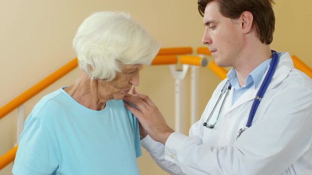 Medium shot of young male physical therapist with stethoscope examining elderly woman suffering from muscle pain