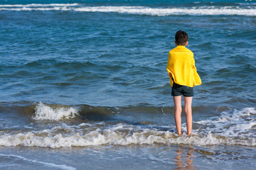 Back view on the boy standing on seashore of the beach in the yellow towel  and looking on the sea....