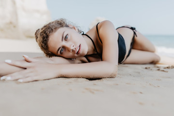 Obraz na płótnie Canvas Happy young woman lying on white sand and looking at camera. Portrait of beautiful woman lying on front at tropical beach. Brunette girl lying on sand in black swimsuit relaxing at sea.