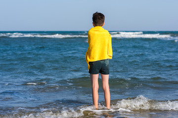 Back view on the boy standing on seashore of the beach in the yellow towel  and looking on the sea. Concept.