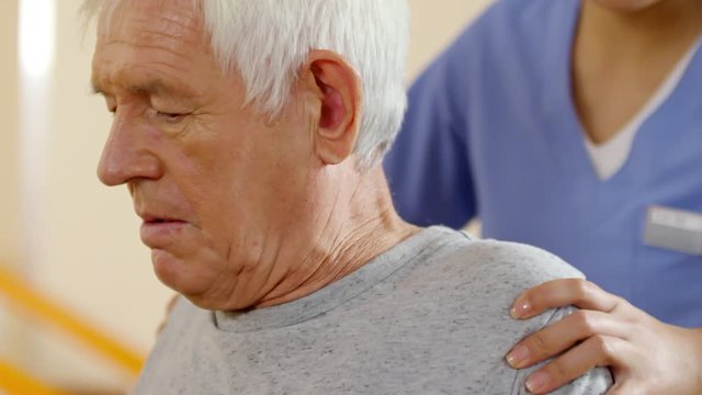 Unrecognizable female physical therapist massaging shoulder and back of senior man with grey hair