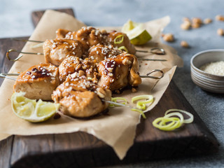 Grilled Asian teriyaki chicken skewers with sesame, leek, peanuts and citrus juice on a paper on wood board on grey background. 
