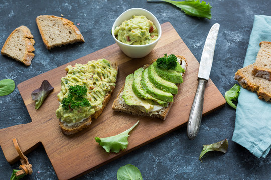 Slices of bread with sliced avocado and avocado cream on wooden board