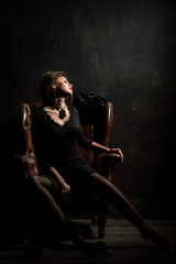 Roaring Twenties. Woman portrait in the style of Gatsby. Low key. A beautiful young woman in a black lace dress is sensually sitting in a brown leather chair.