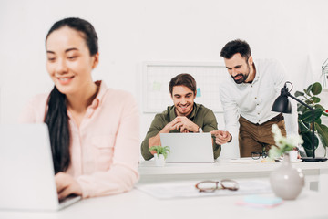 Fototapeta premium Selective focus of male coworkers looking at laptop and smiling in office