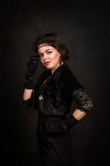Roaring Twenties. Woman portrait in the style of Gatsby. Low key. A beautiful young woman in a black dress touches cheek with the hand on which she wears a black glove.