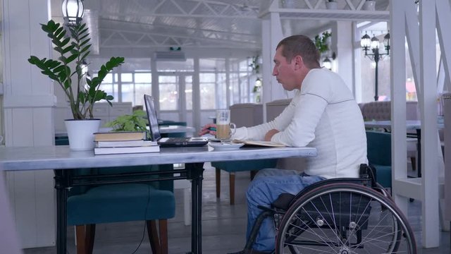 online courses, student crippled man in wheelchair uses a laptop and drinking coffee sitting at a table with books in a cafe