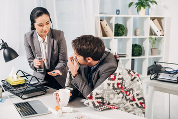 call center operator giving pills and glass of water to sick coworker with cup in office