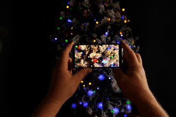 Elderly man holds in his hands new model of smartphone and take pictures of garlands and Christmas lights on New Year tree. Christmas Eve