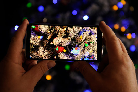 Elderly man holds in his hands new model of  smartphone and take pictures of garlands and Christmas lights on New Year tree. Christmas Eve