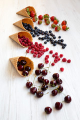 Waffle sweet ice cream cones with raspberries, cherries, strawberries and blueberries over white wooden background, side view.