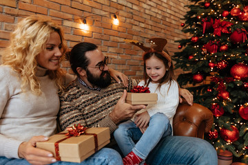 Christmas. Family. Home. Togetherness. Happy parents are giving gifts for their little girl; on the couch near the Xmas tree