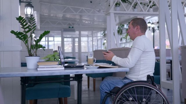 senior handicapped man in wheel chair sitting at a table with cup of coffee talking on a mobile phone and uses a laptop in a restaurant.