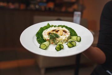 The waiter keeps the dish with Russian salad decorated with mushrooms and fresh cucumbers. Chef holding plate with tasty and delicious food and salad close up.