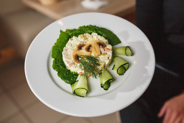 The waiter keeps the dish with Russian salad decorated with mushrooms and fresh cucumbers. Chef holding plate with tasty and delicious food and salad close up.