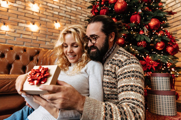 Christmas. Couple. Home. Togetherness. Man is giving a gift to his woman, both are smiling near the Christmas tree