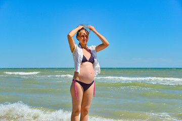 Fototapeta na wymiar A happy and joyful pregnant woman stands by the sea and ocean on the beach under the sun in the open air, hair develops in the wind. Hands holding up and enjoying the sunshine.