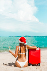 Beautiful  Santa girl in bikini and Christmas hat on the beach of a tropical island, in the hands of a glass of champagne and a red suitcase. Holiday concept for the Christmas holidays and New Year