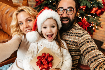 Christmas. Family. Home. Togetherness. Happy parents and their little girl are looking at camera and smiling near the Xmas tree