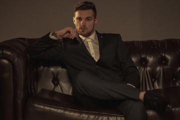 Young businessman in a suit and a shirt is sitting and pondering on the leather sofa.