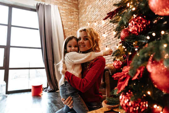 Christmas. Childhood. Home. Little girl and her mom are standing near the Xmas tree, hugging, looking at camera and smiling
