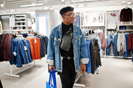 Stylish casual african american man at jeans jacket and black beret with fanny pack or waist bag holding blue shopping bags at clothes store.