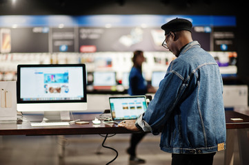 Stylish casual african american man at jeans jacket and black beret using new laptop at electronics...
