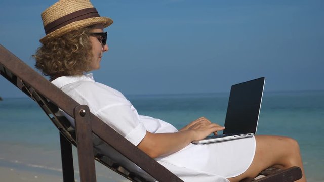Woman Working On Laptop On The Beach By The Sea. Remote Work, Freelance, Digital Nomad Concept.