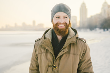 Close-up Caucasian young male red hair and beard in a hat and a park coat posing winter model against a background of a lake to freeze snow. © Elizaveta