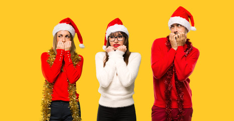 A group of people Blonde woman dressed up for christmas holidays is a little bit nervous and scared putting hands to mouth on yellow background