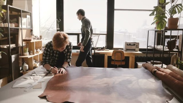 Professional man and woman working with big piece of leather material on a table in modern light manufacturing workshop