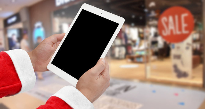 Santa Claus holding white tablet with blank screen, clothing store in background. Christmas shopping concept