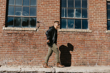 Young Attractive Modern Fashion Caucasian Male Guy Walking, Sitting, Smiling, and Laughing Outside Urban Old Abandoned Brick Warehouse Building in Urban City in Winter Season on Bright Sunny Day