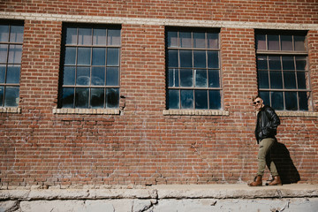 Obraz na płótnie Canvas Young Attractive Modern Fashion Caucasian Male Guy Walking, Sitting, Smiling, and Laughing Outside Urban Old Abandoned Brick Warehouse Building in Urban City in Winter Season on Bright Sunny Day