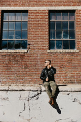 Fototapeta na wymiar Young Attractive Modern Fashion Caucasian Male Guy Walking, Sitting, Smiling, and Laughing Outside Urban Old Abandoned Brick Warehouse Building in Urban City in Winter Season on Bright Sunny Day