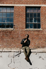 Fototapeta na wymiar Young Attractive Modern Fashion Caucasian Male Guy Walking, Sitting, Smiling, and Laughing Outside Urban Old Abandoned Brick Warehouse Building in Urban City in Winter Season on Bright Sunny Day