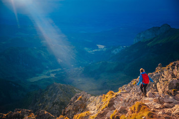 Female tourist with red backpack running in mountains. Orange sunset light in background