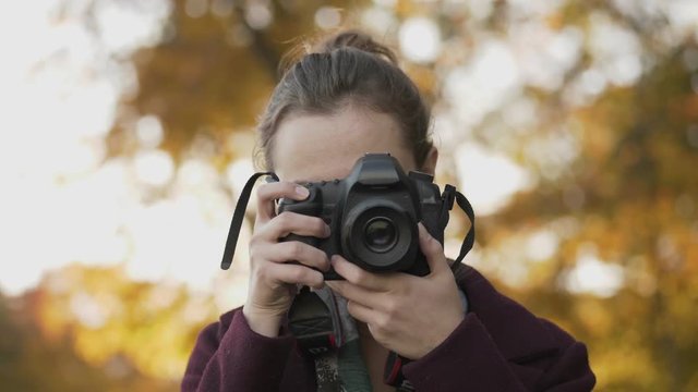 Professional young woman photographer with digital camera taking pictures at autumn city park. Close up, slow motion, shallow depth of field.