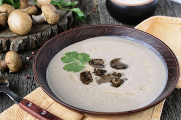 Appetizing mushroom cream soup with pieces of fried mushrooms and cilantro leaf in a dark ceramic bowl on old wooden boards next to whole mushrooms and spoon