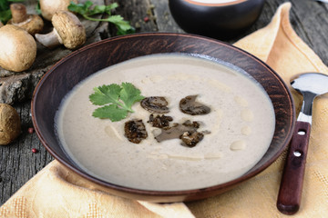 Appetizing mushroom cream soup with pieces of fried mushrooms and cilantro leaf in a dark ceramic bowl on old wooden boards next to whole mushrooms
