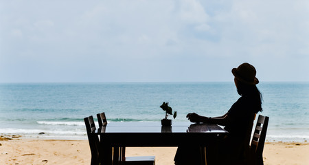 single Asian young woman sits at dining table by window at tropical beach, Lanta island, Krabi, Thailand.