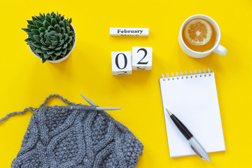 Wooden cubes calendar February 2nd. Cup of tea with lemon, empty open notepad for text. Pot with succulent and gray fabric on knitting needles on yellow background. Top view Flat lay Mockup Concept