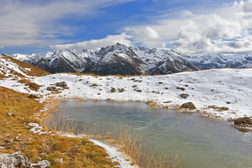 Little frozen pond in the Lechtal Alps surrounded by brown grass in autumn at a sunny day. Snow capped rocky mountains in the background. Tyrol, Austria