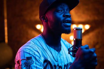 Young man with beard and mustaches in baseball cap and t-shirt sing in microphone. Pink and blue...