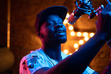 Man with beard and mustaches in baseball cap and t-shirt sing in microphone. Pink and blue neon...