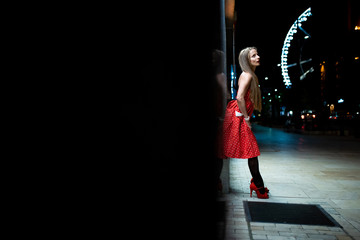 A beautiful mature woman in a red dress standing on the street and leaning to the wall at night...