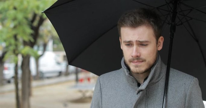 Sad man walking complaining holding an umbrella under the rain in winter in a park
