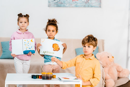 adorable multiethnic children holding pictures and looking at camera while painting together
